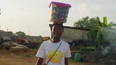 Mujer-Joven-Con-Una-Sonrisa-Llevando-Una-Caja-De-Plástico-En-La-Cabeza-Que-Parece-Unas-Botellas-De-Bebidas-Que-Se-Venden-En-Una-Comunidad-En-Kumasi,-Ghana
