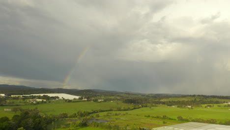 Die-Luftaufnahme-Schwenkt-Sanft-Nach-Links-Und-Zeigt-Eine-Wunderschöne-Naturlandschaft-Unter-Einem-Bunten-Regenbogen-Vor-Dem-Hintergrund-Dichter-Wolken-Am-Himmel