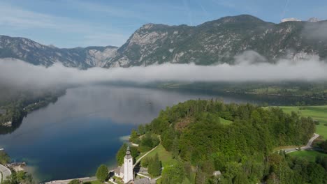 Aerial-view-of-Lake-Bohinj-and-its-church-while-low-clouds-are-entering-the-valley