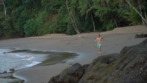 Young-men-running-towards-camera-at-sandy-beach-with-pacific-and-rainforest-in-the-background