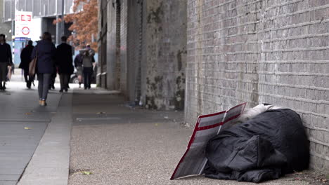 People-walk-past-a-homeless-person-asleep-in-a-grey-sleeping-bag-on-the-floor-against-the-brick-wall-of-an-underpass-during-daylight-in-winter