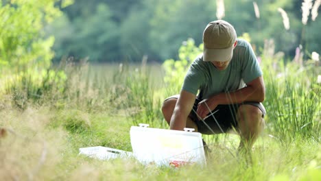 Young-angler-sits-in-high-grass-of-meadow-and-prepares-his-equipment-for-fishing