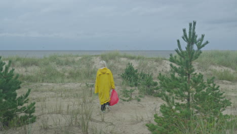Woman-in-yellow-raincoat-with-red-bag-walking-towards-sea-along-sandy-dune,-smooth-tracking-shot