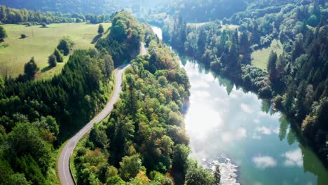 Winding-country-road-along-bright-blue-river-in-which-the-bright-sun-is-reflected-surrounded-by-green-forests,-aerial-drone