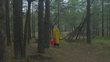 Woman-in-yellow-raincoat-with-red-bag-walking-along-through-forest-toward-sea,-captured-with-a-steady-camera