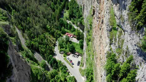 Drone-descends-revealing-a-jagged-rock-face-on-the-right-side-part-of-the-canyon-and-the-parking-space-below-with-buildings-for-visitors-to-come,-Trigrad-Gorge,-Smolyan-Province,-Bulgaria
