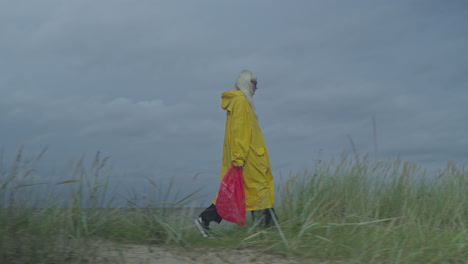 Woman-in-yellow-raincoat-with-red-bag-walking-along-sandy-dune,-captured-with-a-sideways-camera-track
