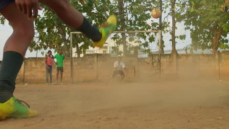 A-boy-tries-to-be-the-goalkeeper-as-this-worn-out-football-is-kicked-towards-the-goal-and-misses-to-the-right,-community-football-pitch,-Kumasi,-Ghana