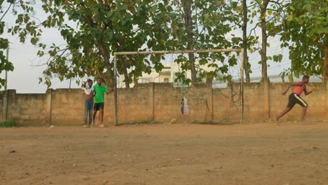 Worn-out-football-kicked-towards-the-goal-as-the-goalkeeper-successfully-stops-it-from-making-a-score-tossing-to-it-to-the-left,-community-soccer-field,-Kumasi,-Ghana