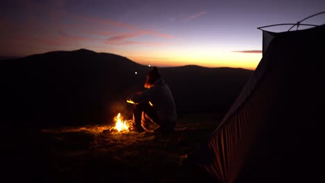 Man-enjoys-bonfire-alone-nature-wilderness-mountain-sunset-tent-camp