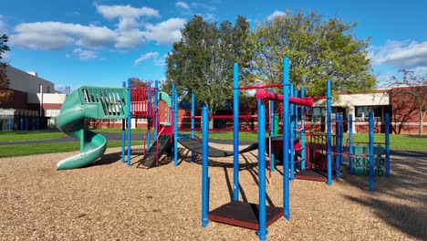 Colorido-Parque-Infantil-Con-Toboganes-Y-Columpios-En-Un-Día-Soleado,-Con-La-Escuela-Al-Fondo-En-EE.UU.