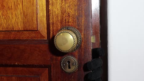 Man-with-a-black-glove-slowly-opens-a-wooden-door-turning-the-door-knob-and-showing-his-hand-and-masked-face