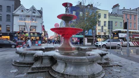 Street-in-Cork-city-with-red-lit-water-fountain,-pedestrians,-public-transportation,-traffic,-evening-time-in-winter-with-Christmas-decorations