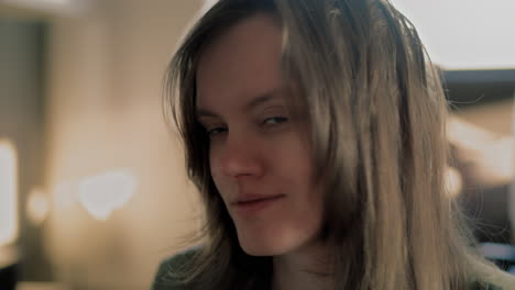 Cinematic-close-up-of-a-Young-White-Woman-looking-straight-into-the-Camera-and-smiling-during-a-golden-hour-Sunrise-in-her-apartment-in-slow-motion
