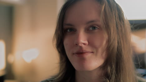 Cinematic-close-up-shot-of-a-beautiful-white-woman's-Face-in-her-apartment-looking-around-and-then-staring-straight-into-the-camera-and-smiling-during-a-golden-hour-sunrise-in-slow-motion