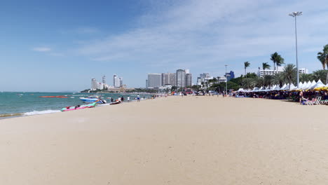 Tall-buildings-in-the-horizon,-hotels,-condominiums,-as-people-walkabout-on-the-beach-in-Pattaya-while-other-choose-to-stay-under-huge-beach-umbrellas-and-canopies,-Thailand