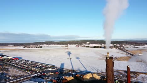 Smoke-stack-emits-grey-white-cloud-of-fumes,-long-shadow-cast-onto-snow-behind-in-industrial-region