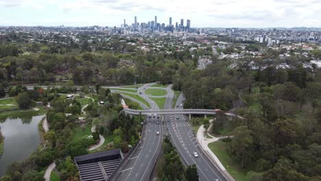 Aerial-view-of-a-major-freeway-with-a-roundabout,-city-skyline-in-the-background-and-a-park-with-ponds-near-by