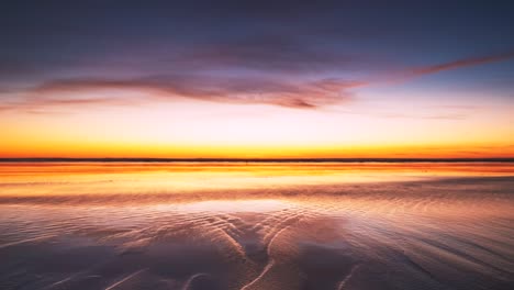 Vibrant-sunset-reflecting-on-the-wet-sand-and-shadows-creeping-in-on-the-timelapse-at-the-end