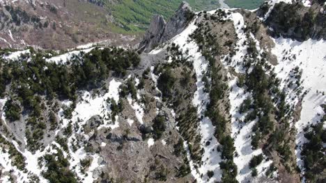 Drone-view-in-Albania-flying-in-the-alps-showing-a-snowy-and-rocky-mountain-peak-between-two-green-forest-valleys-in-Theth