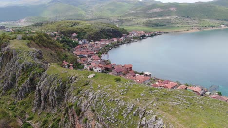 Drohnenansicht-In-Albanien,-Die-über-Eine-Kleine-Stadt-Mit-Häusern-Neben-Einem-Grünen-Hügel-Am-Ohridsee-Mit-Kristallblauem-Wasser-Fliegt