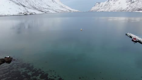 Drone-view-in-Tromso-area-in-winter-flying-over-a-fjord-surrounded-by-white-snowy-mountains-and-pebble-beach-in-Norway