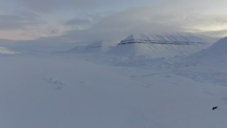 Drone-view-in-Svalbard-panning-over-snowy-white-mountains-with-a-line-of-snowmobiles-in-Norway