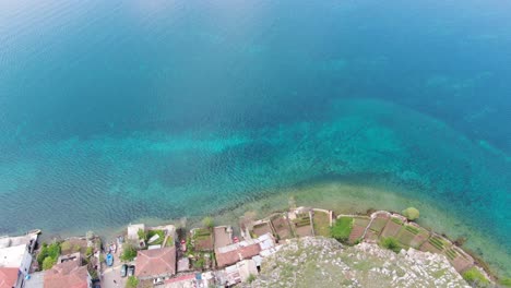 Drone-view-in-Albania-flying-over-a-green-mountain-with-a-small-town-to-Ohrid-lake-top-view-crystal-clear-water