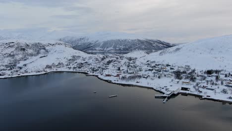 Vista-De-Drones-En-La-Zona-De-Tromso-En-Invierno-Volando-Sobre-Un-Fiordo-Hacia-Un-Pequeño-Pueblo-Nevado-Con-Pequeñas-Casas-En-Noruega