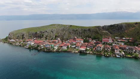 Drohnenansicht-In-Albanien,-Die-über-Eine-Kleine-Stadt-Mit-Häusern-Neben-Einem-Grünen-Hügel-Am-Ohridsee-Mit-Kristallblauem-Wasser-Fliegt