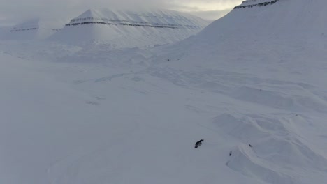 Drone-view-in-Svalbard-flying-over-snowy-white-valley-with-a-line-of-snowmobiles-surrounded-by-mountains-in-Norway