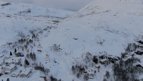 Vista-De-Drones-En-La-Zona-De-Tromso-En-Invierno-Volando-Sobre-Un-Pequeño-Pueblo-Con-Un-Paisaje-Lleno-De-Nieve-Y-Montañas-Blancas-Junto-A-Un-Fiordo-En-Noruega