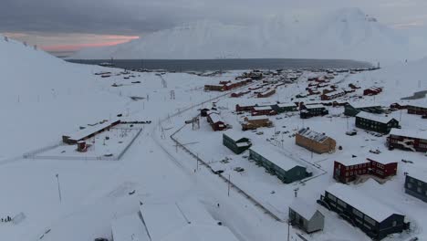 Drone-view-in-Svalbard-flying-over-Longyearbyen-town-showing-houses-in-a-snowy-area-with-a-fjord-and-mountain-in-Norway
