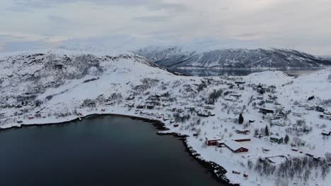 Drone-view-in-Tromso-area-in-winter-flying-over-a-fjord-towards-a-small-snowy-town-with-small-houses-in-Norway