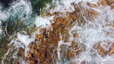 Top-view,-powerful-waves-washing-over-brown-rocky-shore,-drone-stable-shot