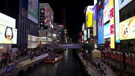Blick-Auf-Touristen-Von-Der-Brücke-Aus,-Die-Nachts-Am-Dotonbori-Kanal-Entlang-Mit-Neonbeleuchteten-Werbetafeln-In-Osaka-Spazieren