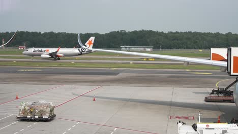 Jetstar-Plane-Taxiing-On-Runway-In-Background-Behind-View-Of-Wing-Of-Plane-Parked-At-Terminal-Gate-At-Changi-Airport-In-Singapore