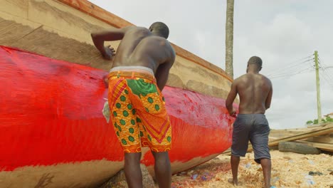 Equipo-De-Dos-Trabajadores-Pintando-Un-Barco-Tradicional-Africano-De-Pescadores-De-Madera-Como-Mantenimiento-De-Rutina.