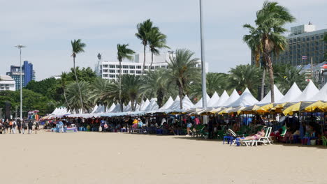 Busy-beach-with-people-under-canopies-and-umbrellas-while-others-walk-around-sunbathing-during-a-windy-day,-Pattaya,-Thailand
