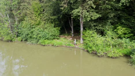 Male-angler-stands-on-shore-of-cloudy-lake-between-grass-and-trees-catching-fish-with-fishing-rod,-drone-panoramic-aerial