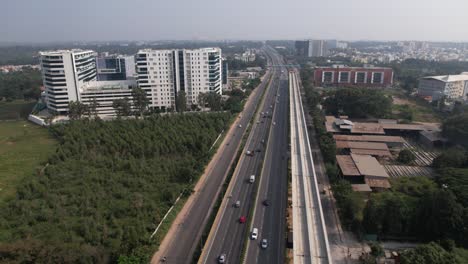 Aerial-footage-of-an-Indian-highway-that-is-expanding-quickly,-with-a-service-road,-fast-moving-automobiles,-and-a-metro-train-bridge-under-construction