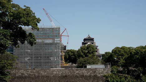 Der-Uto-Turm-Ist-Mit-Gerüsten-Bedeckt-Und-Ein-Baukran-Für-Renovierungsarbeiten-Mit-Der-Burg-Kumamoto-Im-Hintergrund-Hinter-Bäumen-An-Einem-Klaren,-Sonnigen-Tag
