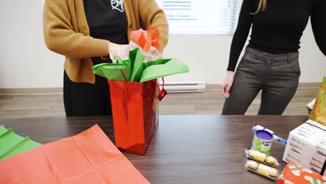 Slow-Motion-4K-Two-Young-Attractive-Caucasian-Woman-Finish-Wrapping-a-Red-Holiday-Colorful-Bag-Christmas-Eve-Decorative-Gift-Present-for-Corporate-Office-Secret-Santa-Festivities