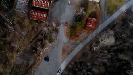 Drone-top-shot-of-a-car-passing-by-on-a-gravel-road-at-winter-near-some-sheds-with-rusty-roofs