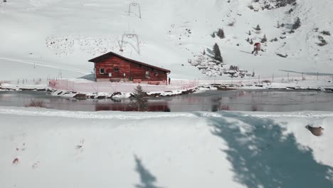 Retreating-drone-shot-of-a-cabin-beside-a-man-made-lake,-and-tilting-to-show-the-pine-trees-on-its-slope,-in-Engelberg-Brunni,-Bahnen-in-Switzerland