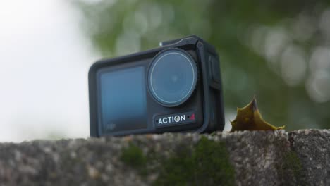 Low-Angle-Produktaufnahme-Der-DJI-Osmo-Action-4-Kamera,-Die-Auf-Einer-Steinmauer-Mit-Grünem-Bokeh-Baumhintergrund-Ruht