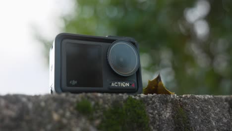 Low-Angle-Product-Shot-Of-DJI-Osmo-Action-4-Camera-Resting-On-Stone-Wall-With-Green-Bokeh-Tree-Background