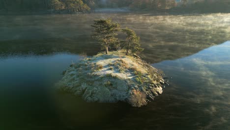 Small-island-with-trees-in-calm-lake-with-mist-flowing-at-sunrise-in-autumn