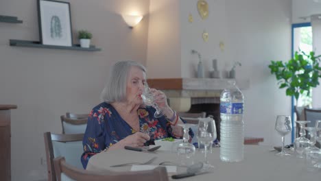 Senior-woman-sitting-alone-at-empty-dining-table,-drinking-water-out-of-glass,-modern-indoor-retirement-home