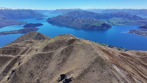 Flyover-famous-Roys-Peak-hiking-track-mountain-summit,-wide-view-of-Lake-Wanaka,-New-Zealand